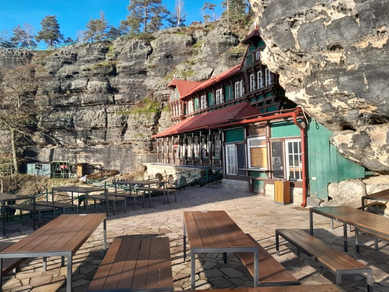 A rustic building with a red roof nestled against a rocky cliff, outdoor tables in the sunny foreground.