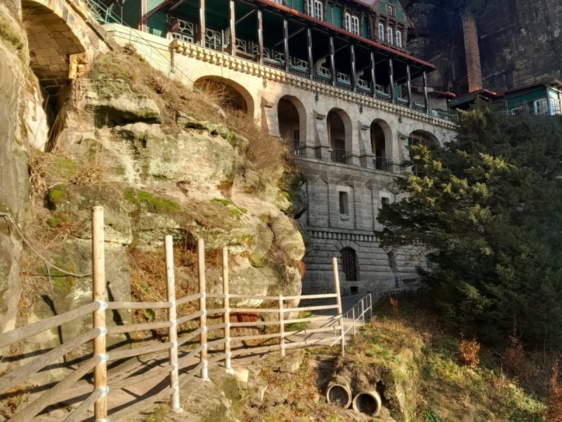 A historic wooden building with arches on a rocky cliff, surrounded by trees and a blue sky.