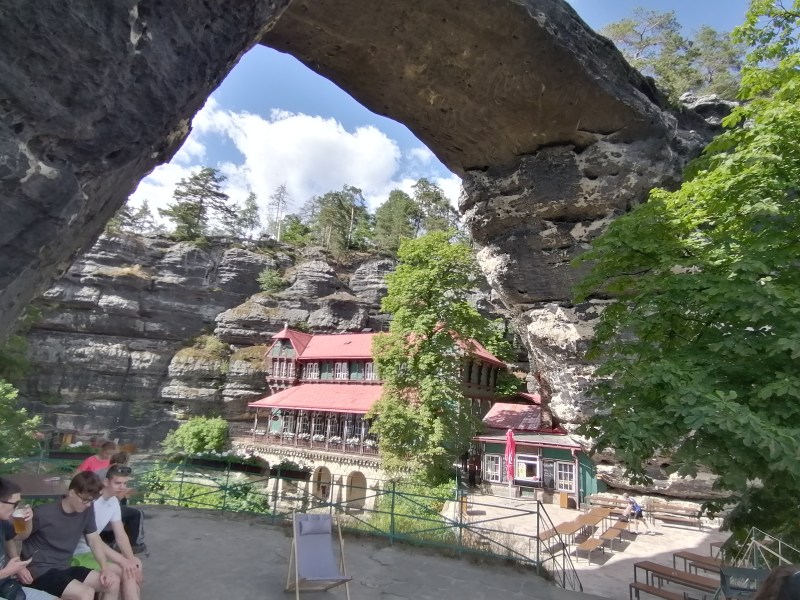 Group seated near stone arch, overlooking a rustic building with red roof, surrounded by trees and rocky cliffs.