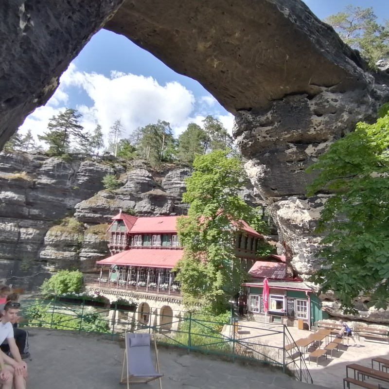 Group seated near stone arch, overlooking a rustic building with red roof, surrounded by trees and rocky cliffs.