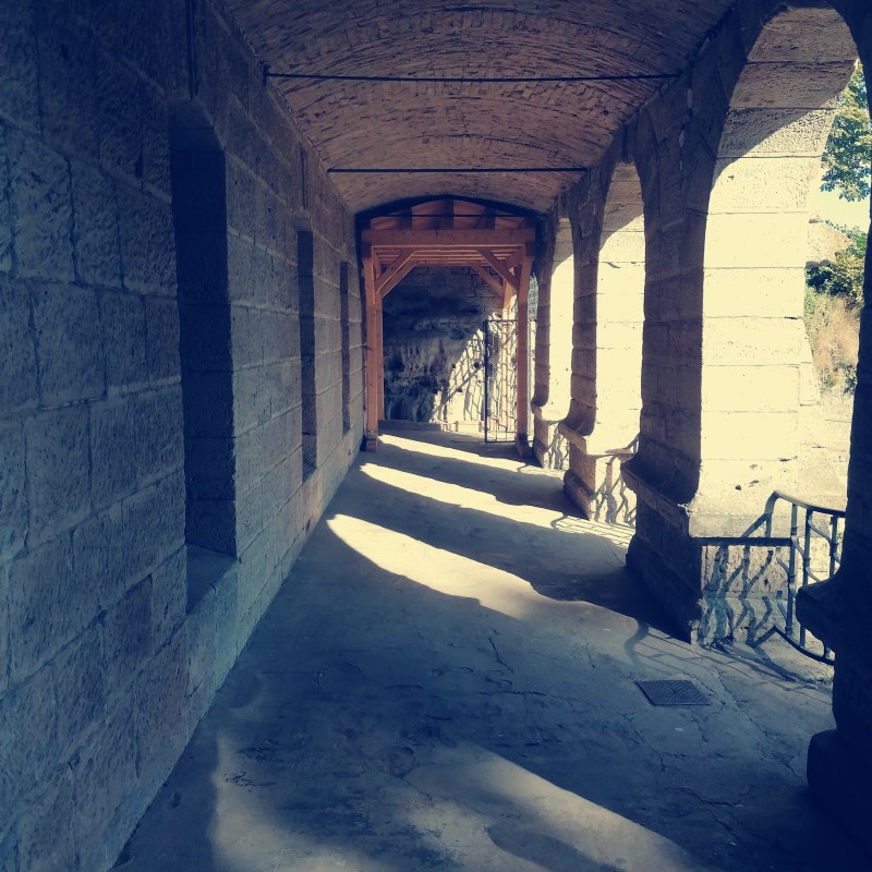 Stone corridor with arches and shadows on the ground, leading to a wooden gate.