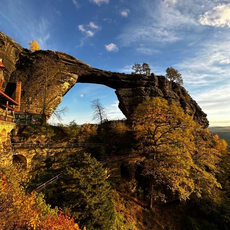 Natural rock arch bridge over forest with autumn foliage under blue sky.