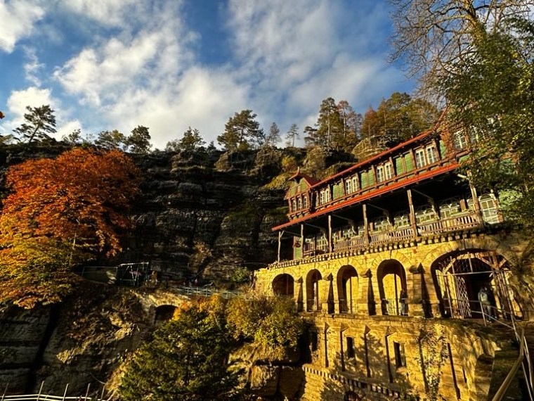 Historic building on a cliffside with arched windows, surrounded by trees under a blue sky with clouds.