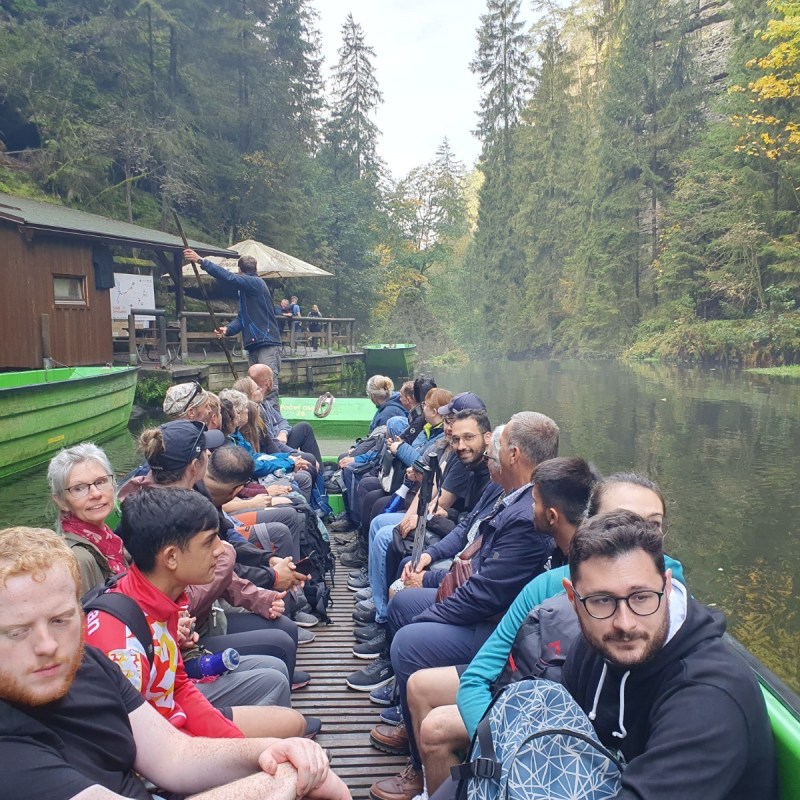 People sitting in a boat on a narrow forested river, wooden cabin on the left.
