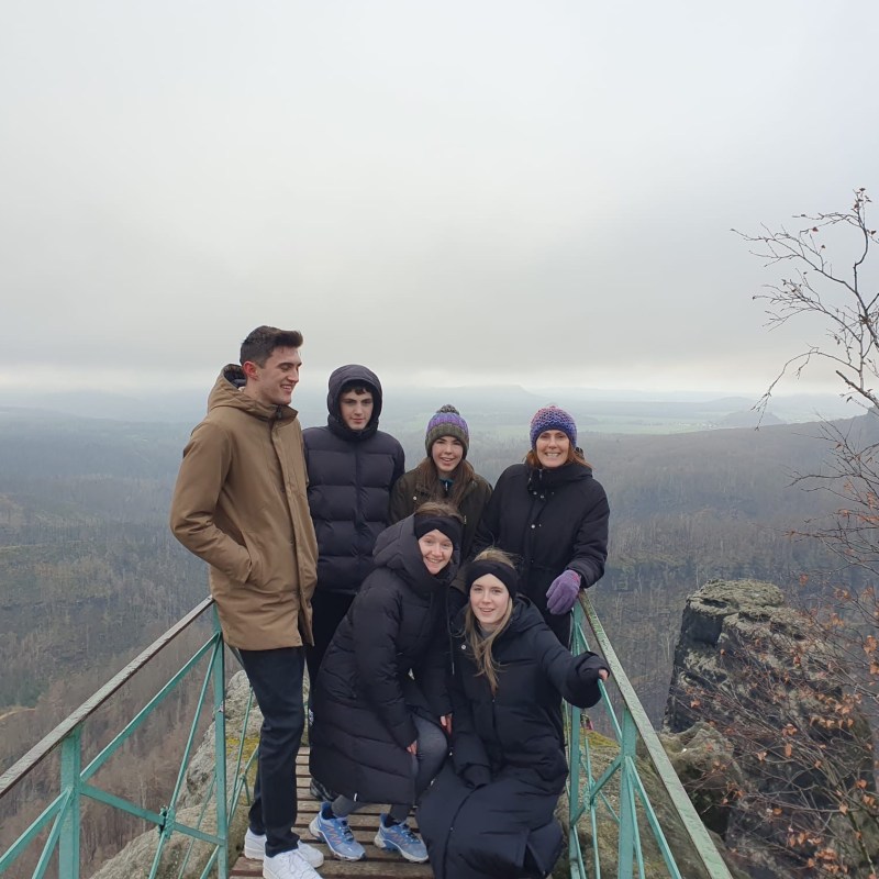 Group of six people on a mountain viewing platform in winter clothing, with a foggy landscape behind.