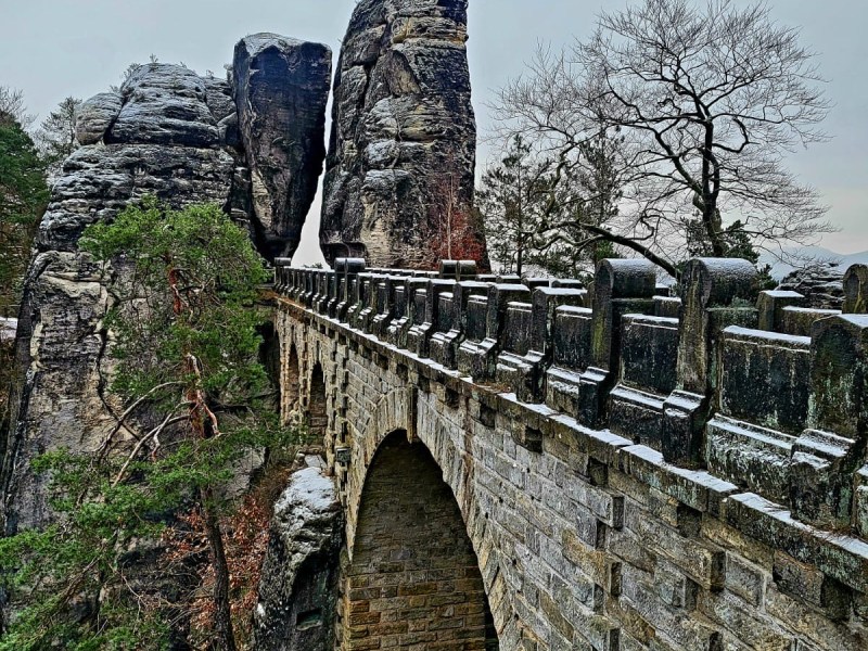Stone bridge with arches, flanked by tall rocks and bare trees in a winter landscape.
