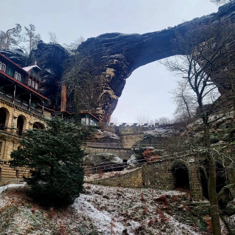 Snow-covered stone arch with adjacent building and bare trees in a winter landscape.
