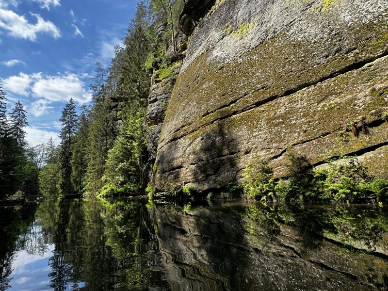 Rocky cliff and trees reflected in a calm river under a blue sky with clouds.