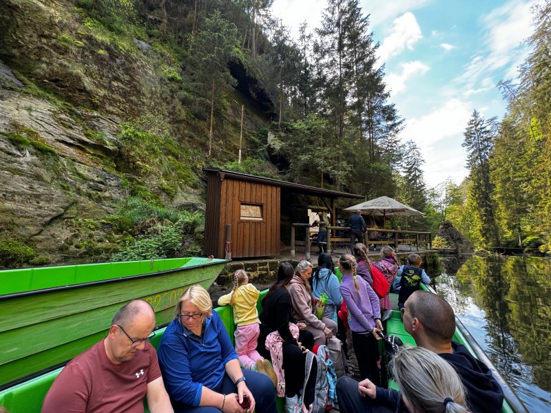 People on a green boat in a forest river, passing a wooden hut.