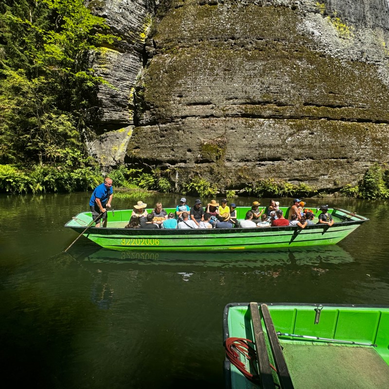 A green boat with people glides through water near a rocky cliff, surrounded by lush greenery.