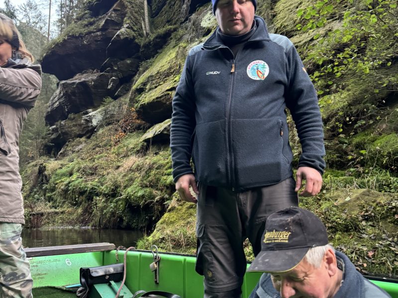 Two men in outdoor gear on a green boat with rocky forest background.