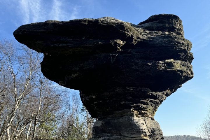 Large balancing rock formation against a clear blue sky with trees in background.