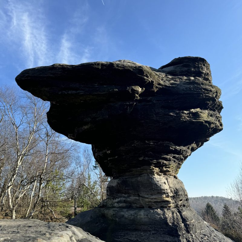 Large balancing rock formation against a clear blue sky with trees in background.