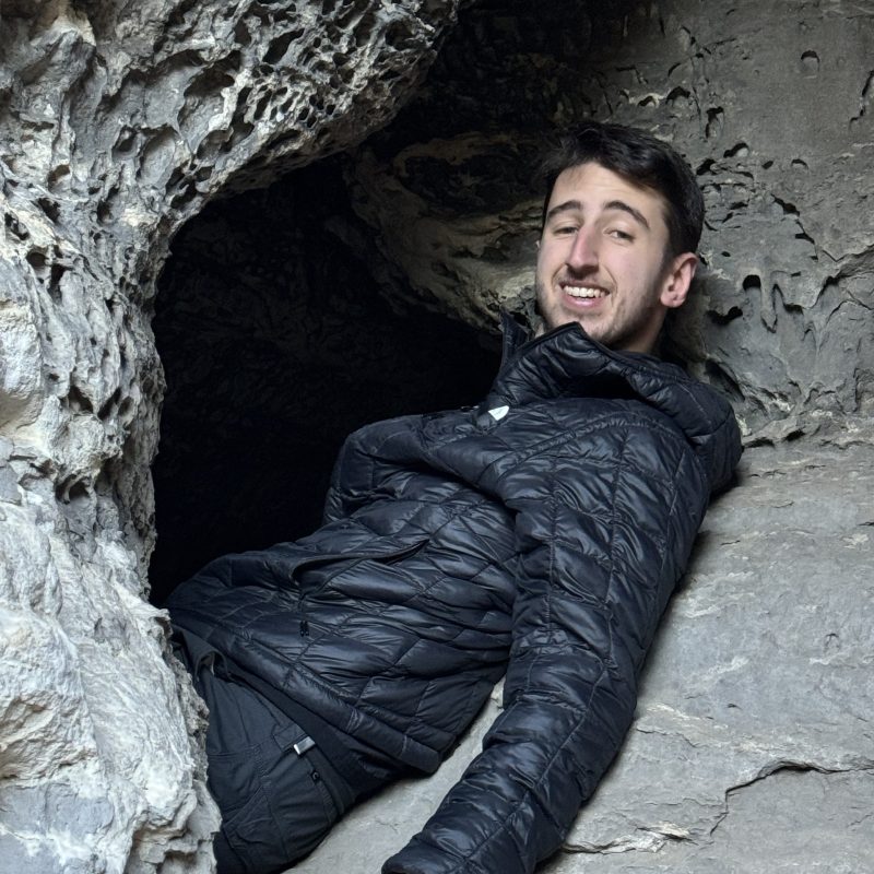 Person in black jacket smiling while reclining in a rocky cave entrance.