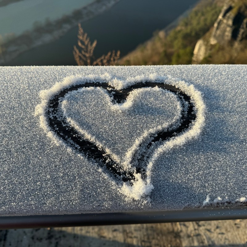 Heart shape drawn on frosty surface overlooking a river and trees.