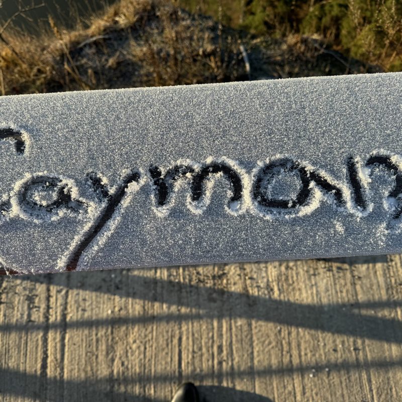 Snow-covered surface with the word 'Cayman' written in it, shadow and grass visible.