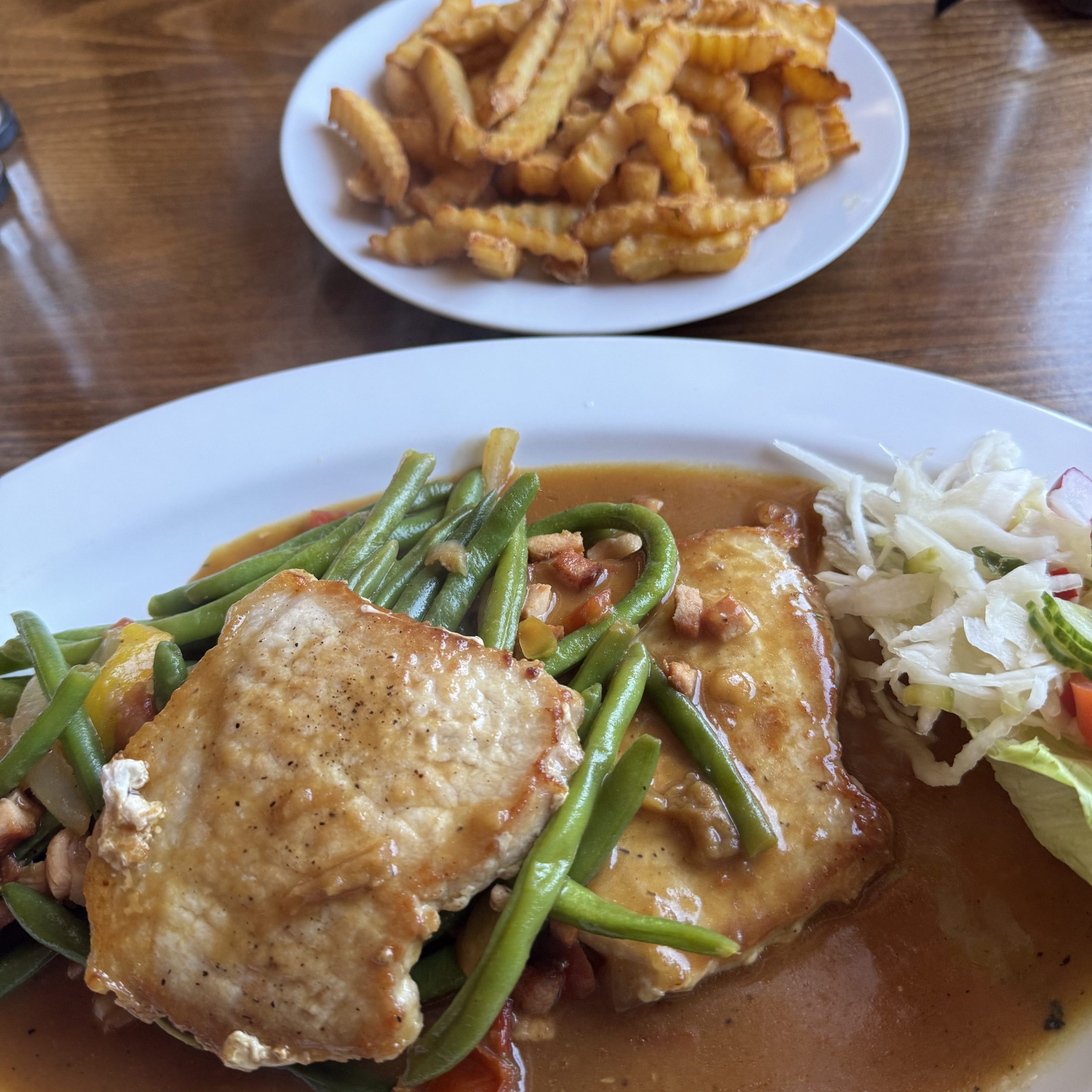 Dish with pork, green beans, sauce, and salad. Plate of crinkle-cut fries in the background.
