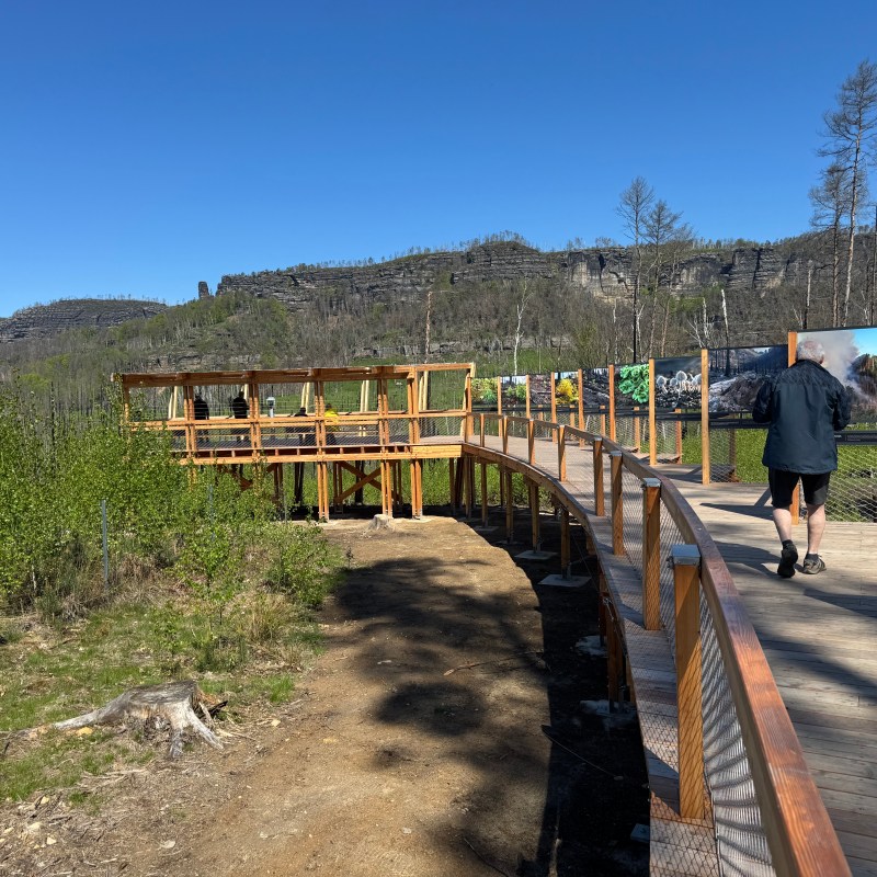 Person walking on a wooden boardwalk with scenic landscape and photo displays.
