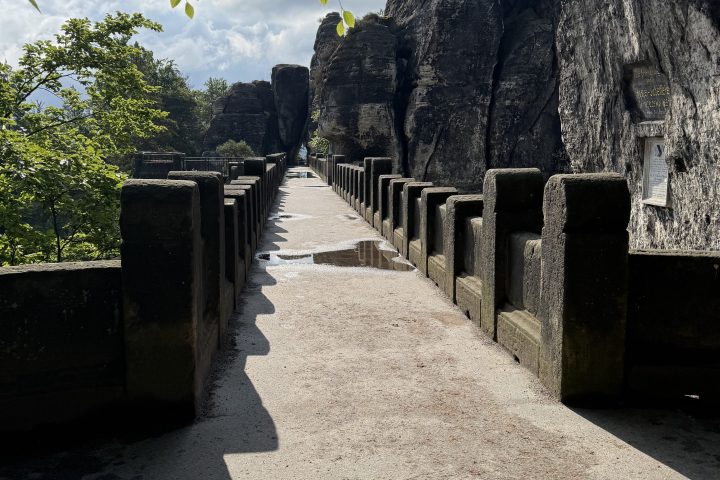 Stone bridge leading to tall rock formations, surrounded by trees under a cloudy sky.