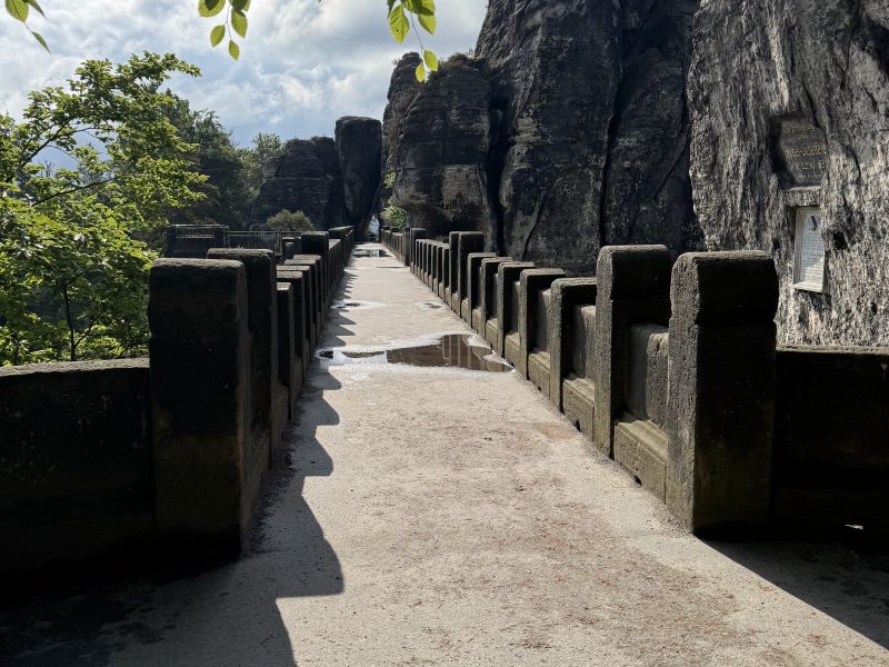Stone bridge leading to tall rock formations, surrounded by trees under a cloudy sky.