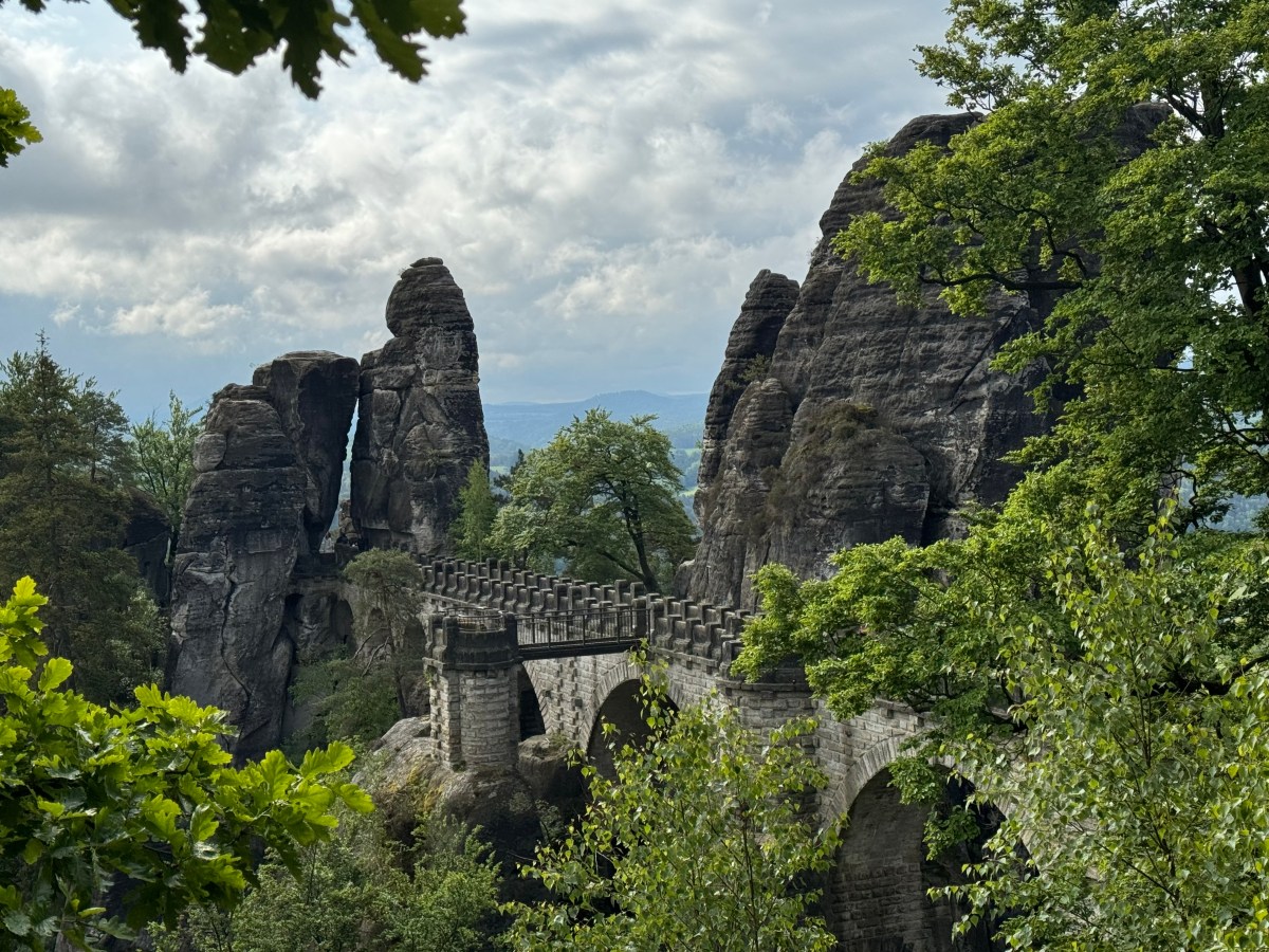 IMG_7064 Stone bridge surrounded by lush green trees and tall rock formations under a cloudy sky.