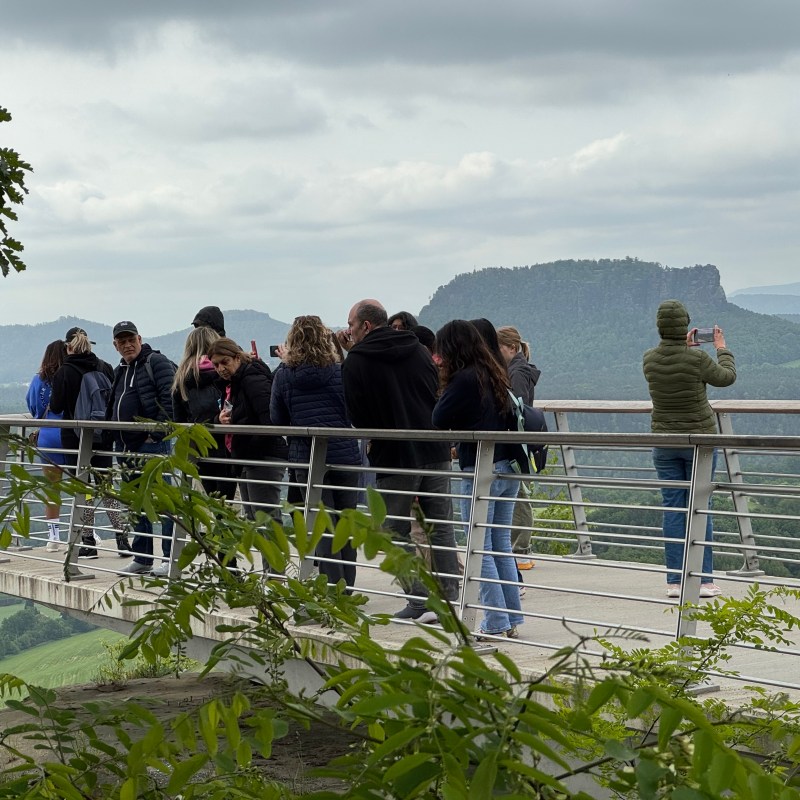 People on a viewing platform overlooking a landscape with hills and cloudy sky.
