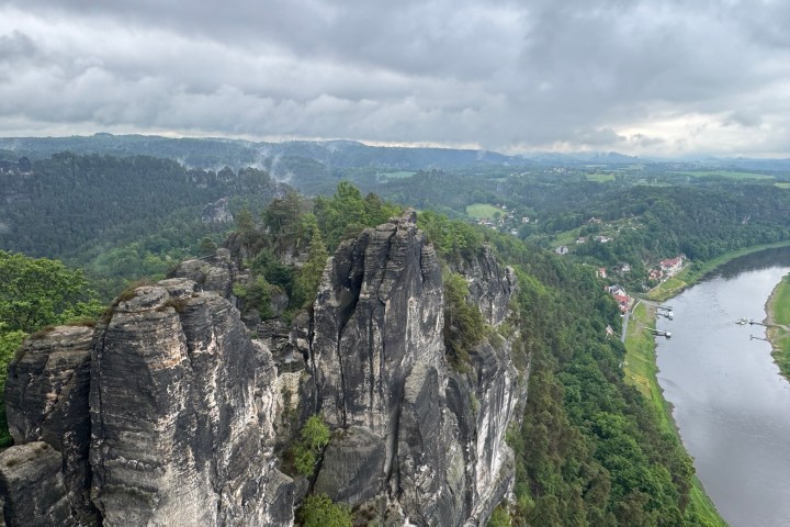 Misty cliffside with green trees and river below, cloudy sky above.