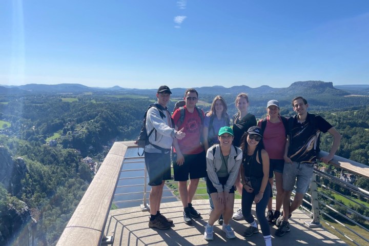 Group of people posing on a scenic viewpoint with mountains and forest in the background under a clear blue sky.