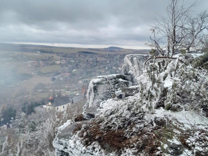 Snow-covered rocky cliff with distant town under cloudy sky.