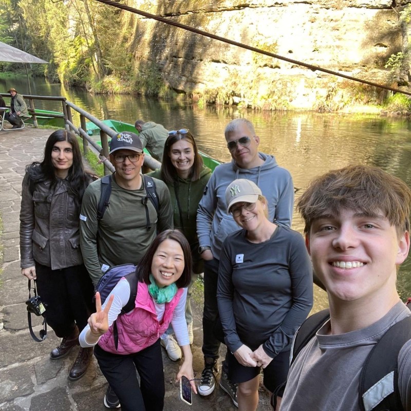 Group of seven people posing near a river with a rocky background on a sunny day.