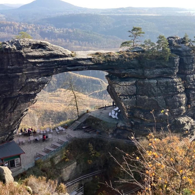 Rock arch formation with a small building and trees in a mountainous landscape.