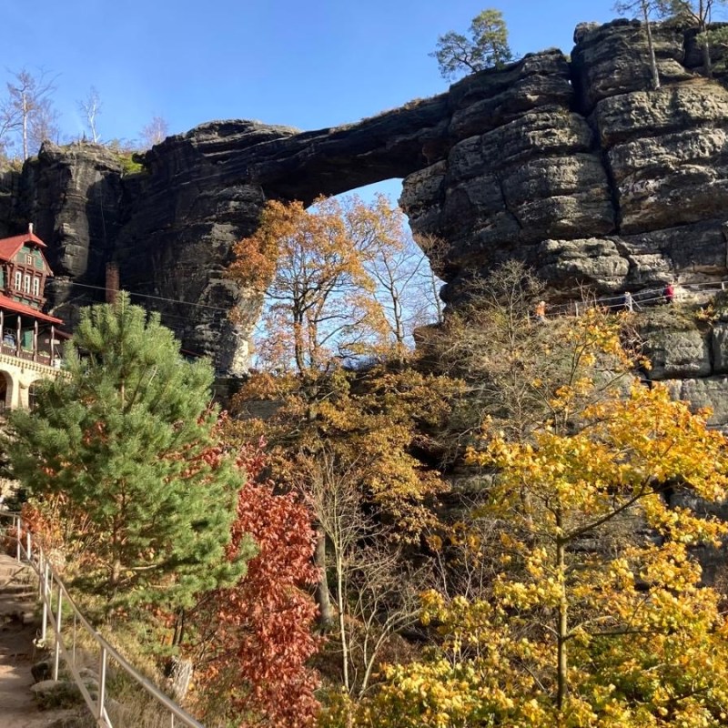 Rock bridge over trees and hikers on path near rustic building in rocky landscape.
