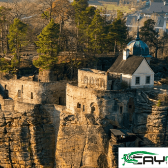 Aerial view of an ancient stone fortress atop a rocky cliff surrounded by trees, with a travel logo overlay.