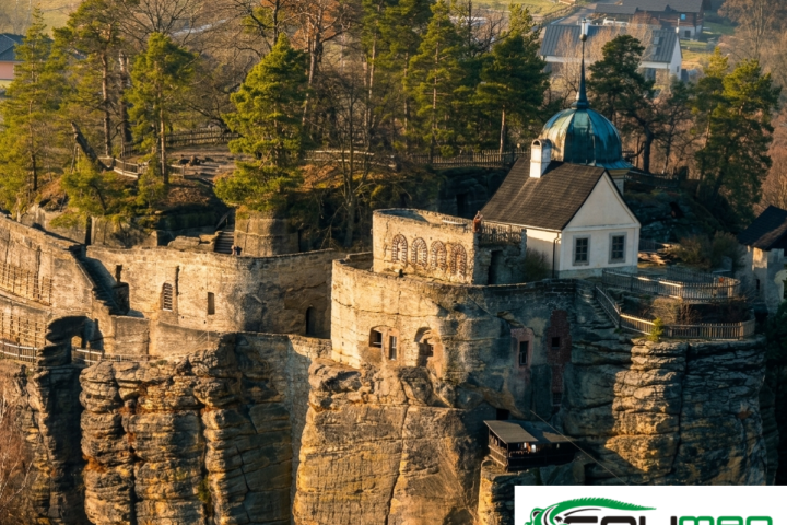 Aerial view of an ancient stone fortress atop a rocky cliff surrounded by trees, with a travel logo overlay.