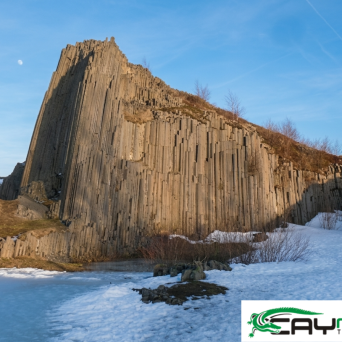 Tall, columnar basalt rock formation with snow at its base under a clear blue sky.