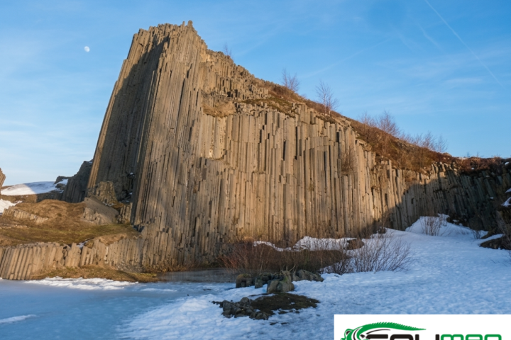 Tall, columnar basalt rock formation with snow at its base under a clear blue sky.