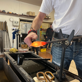 Person shaping molten glass with a tool in a workshop setting.