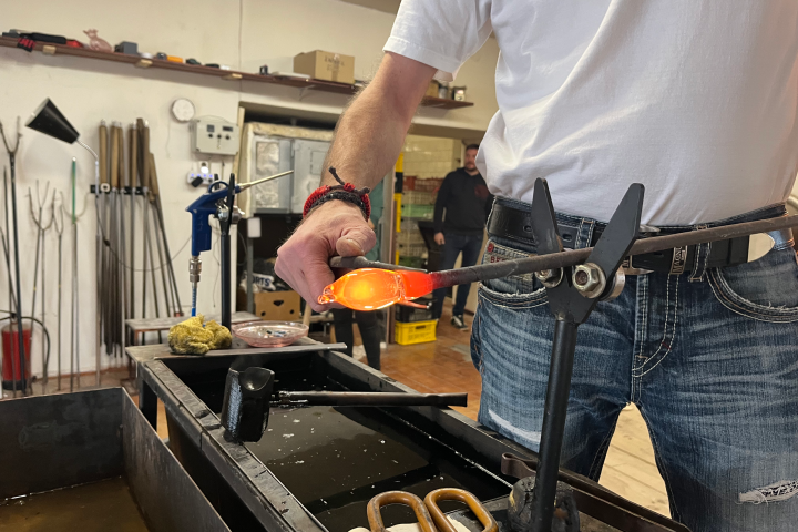 Person shaping molten glass with a tool in a workshop setting.