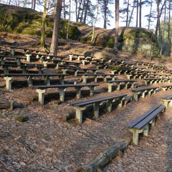 Outdoor amphitheater with wooden benches surrounded by trees and rocky terrain.