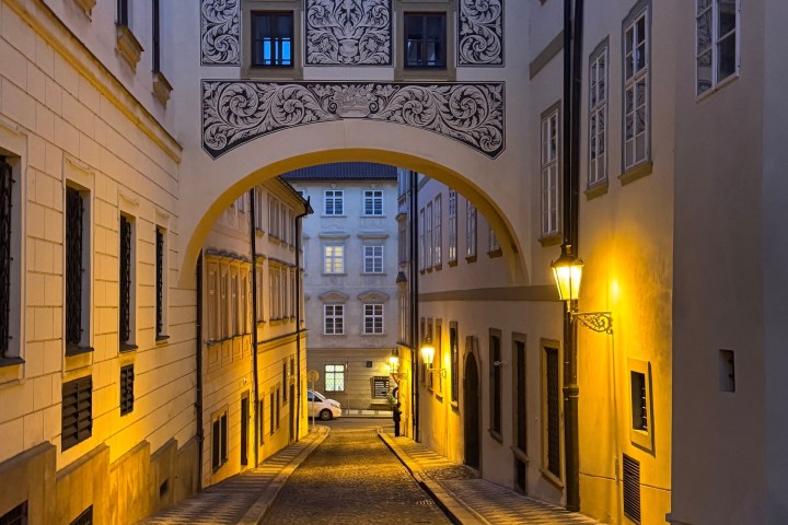 Narrow cobblestone street with archway and ornate designs, lit by street lamps at dusk.