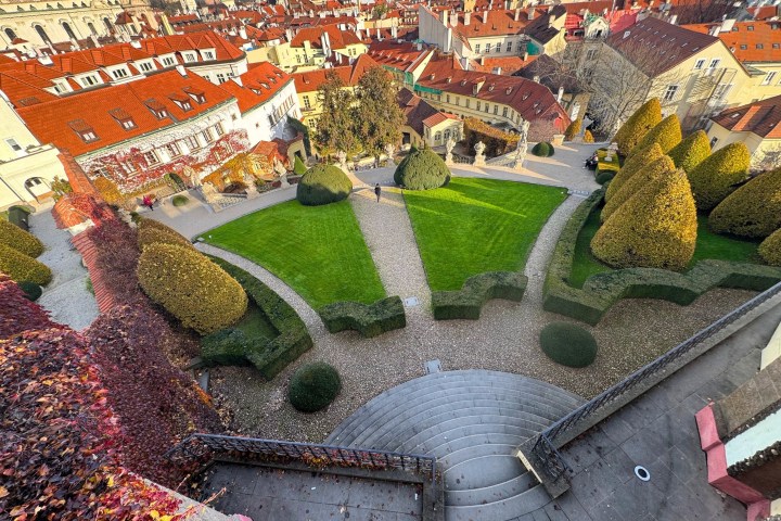 Aerial view of a neatly landscaped garden with grass, shrubs, and circular paths, surrounded by red-roofed buildings.