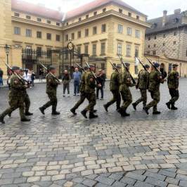 Soldiers in uniform marching in a cobblestone square with onlookers nearby.