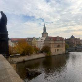 Historic bridge view with statues, river, and old buildings under cloudy sky.